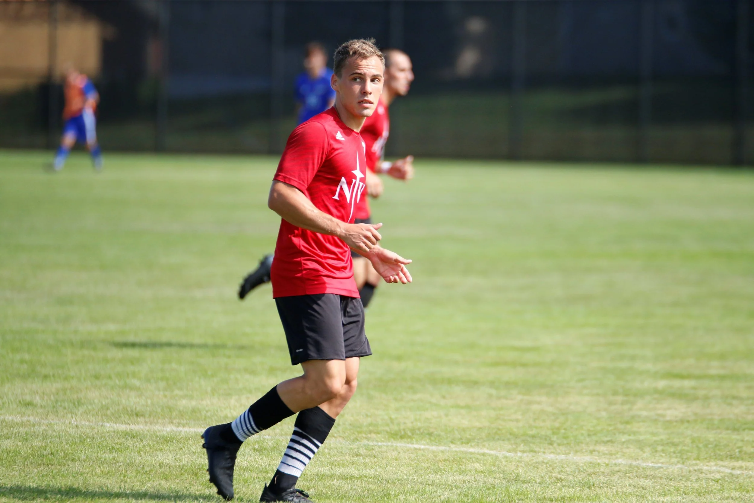 Un jugador de fútbol en un campo de césped, vistiendo camiseta roja, pantalones cortos negros y calcetines con rayas. Parece estar en medio de un partido con otros jugadores en el fondo.