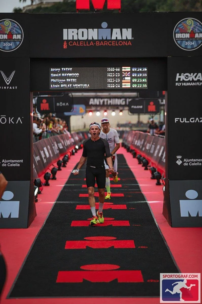 Atleta cruzando la meta en el Ironman Calella-Barcelona, con una expresión de triunfo, en un recorrido con un tapete negro y letras rojas, rodeado de espectadores y anuncios deportivos.