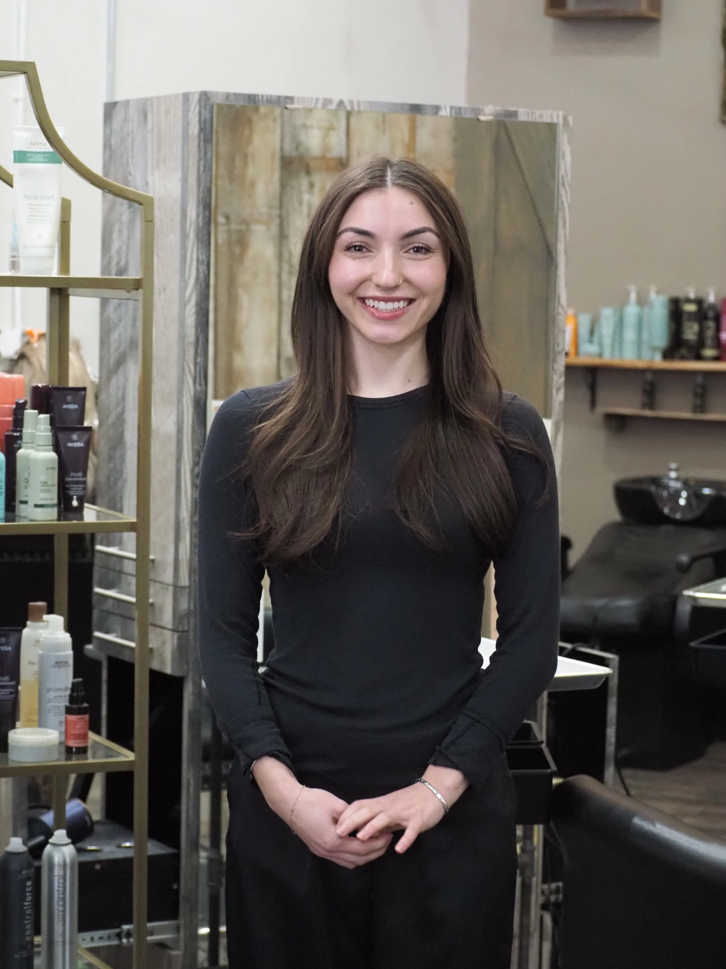 woman in black shirt and dark hair standing in a salon as a salon assistant.