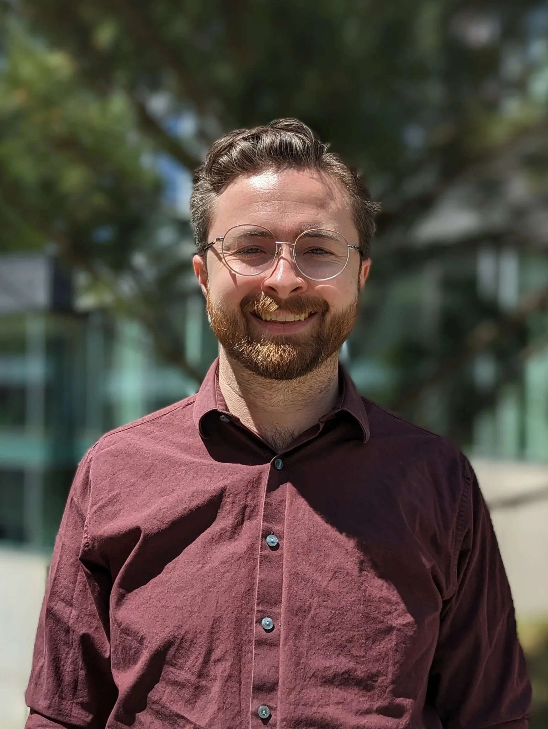 A man with short brown hair and glasses, wearing a maroon shirt in front of a tree and building.