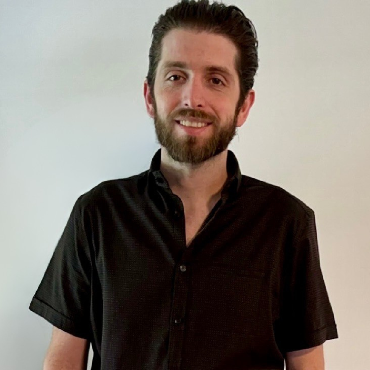 A man with a beard and mustache, wearing a black button-up shirt, smiling and standing against a plain white background.