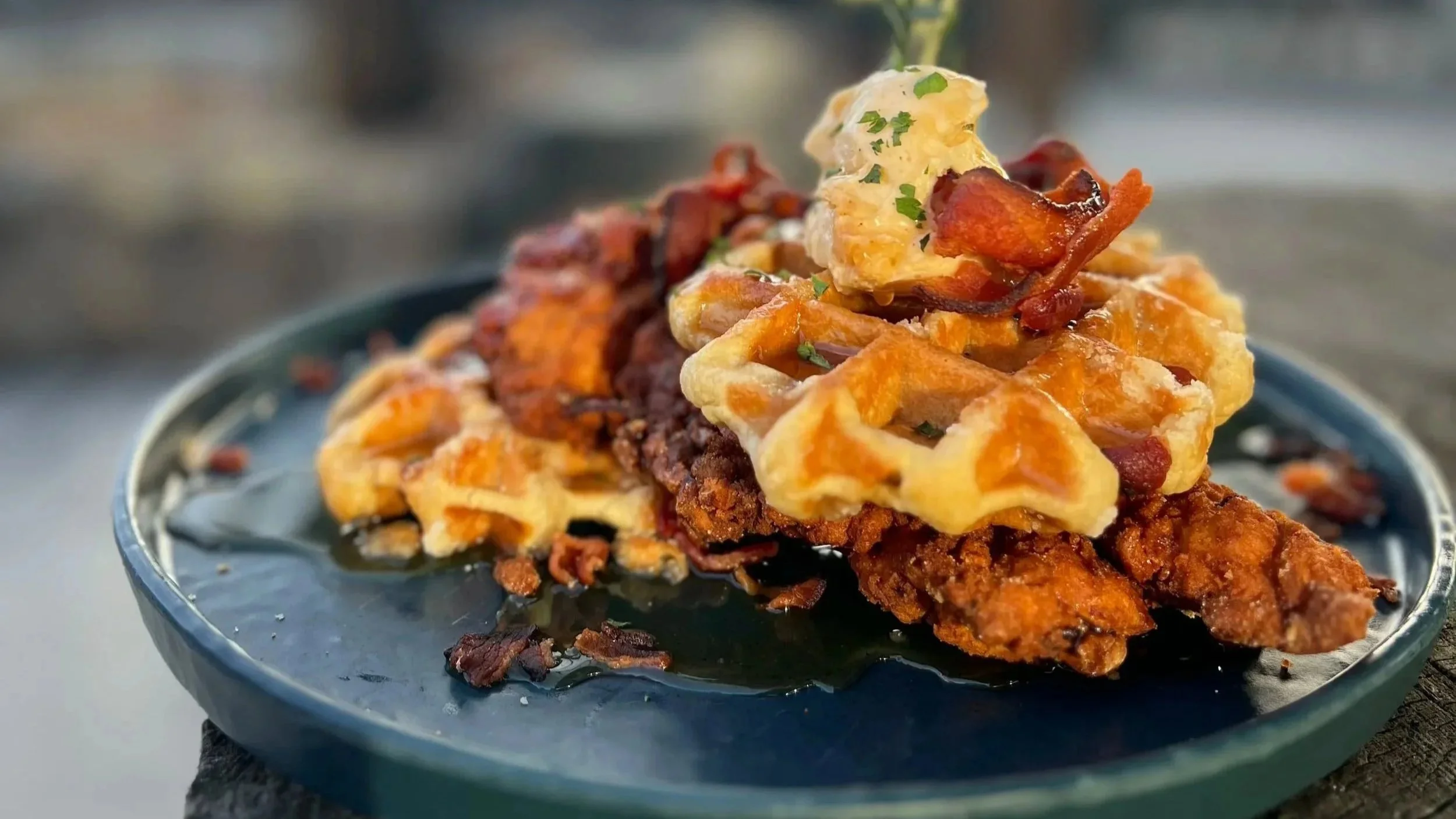 Chicken and waffles with bacon, garnished with herbs on a dark plate.