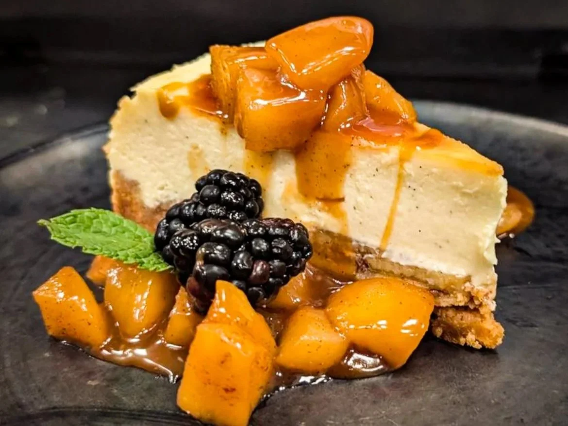 Slice of cheesecake with caramelized fruit topping, blackberries, and a mint leaf, served on a dark plate.