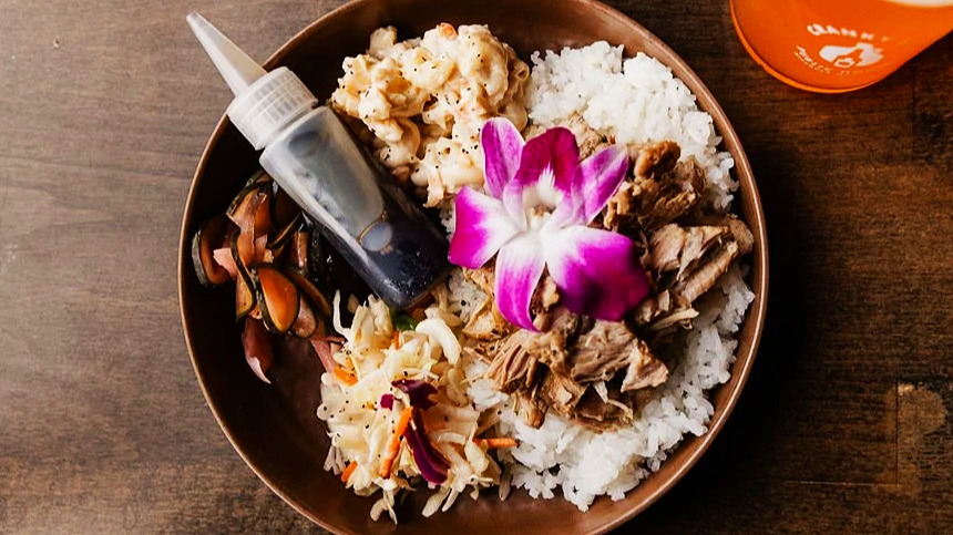 A bowl of Hawaiian plate lunch with rice, macaroni salad, shredded pork with an orchid flower garnish, and a side of vegetables, with a small soy sauce bottle on the side.