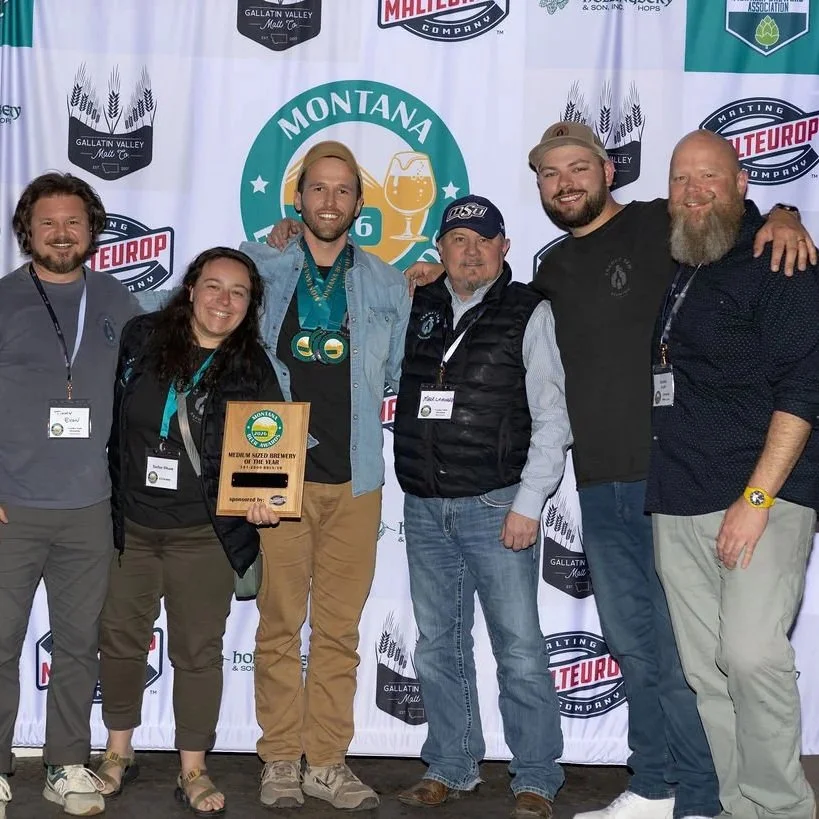 Group of six people standing together at a beer brewing event, with a backdrop featuring logos of Gallatin Valley Malting, Malteurop, and Montana. One person is holding a plaque.