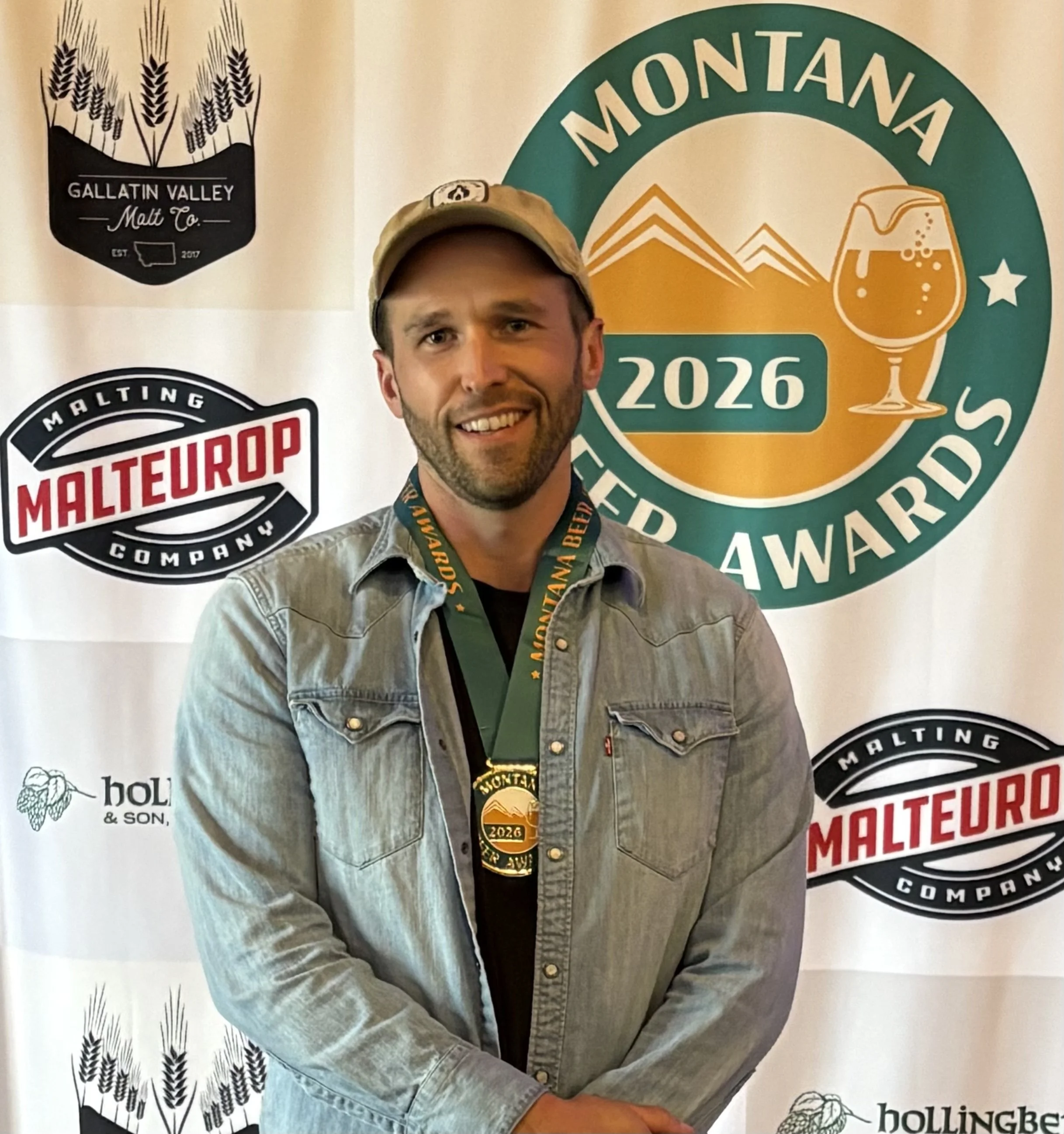 A man standing in front of a backdrop with logos and text, wearing a medal around his neck and a tan baseball cap, smiling at the camera.