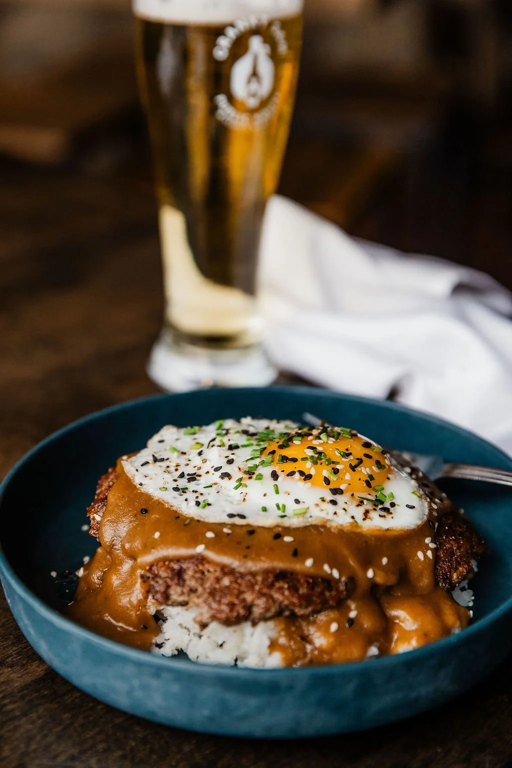 Loco moco, hawaiian street food classic. A plate with a burger patty topped with a fried egg, sesame seeds, chopped chives, and black sesame seeds, accompanied by a glass of beer on a dark wooden table.