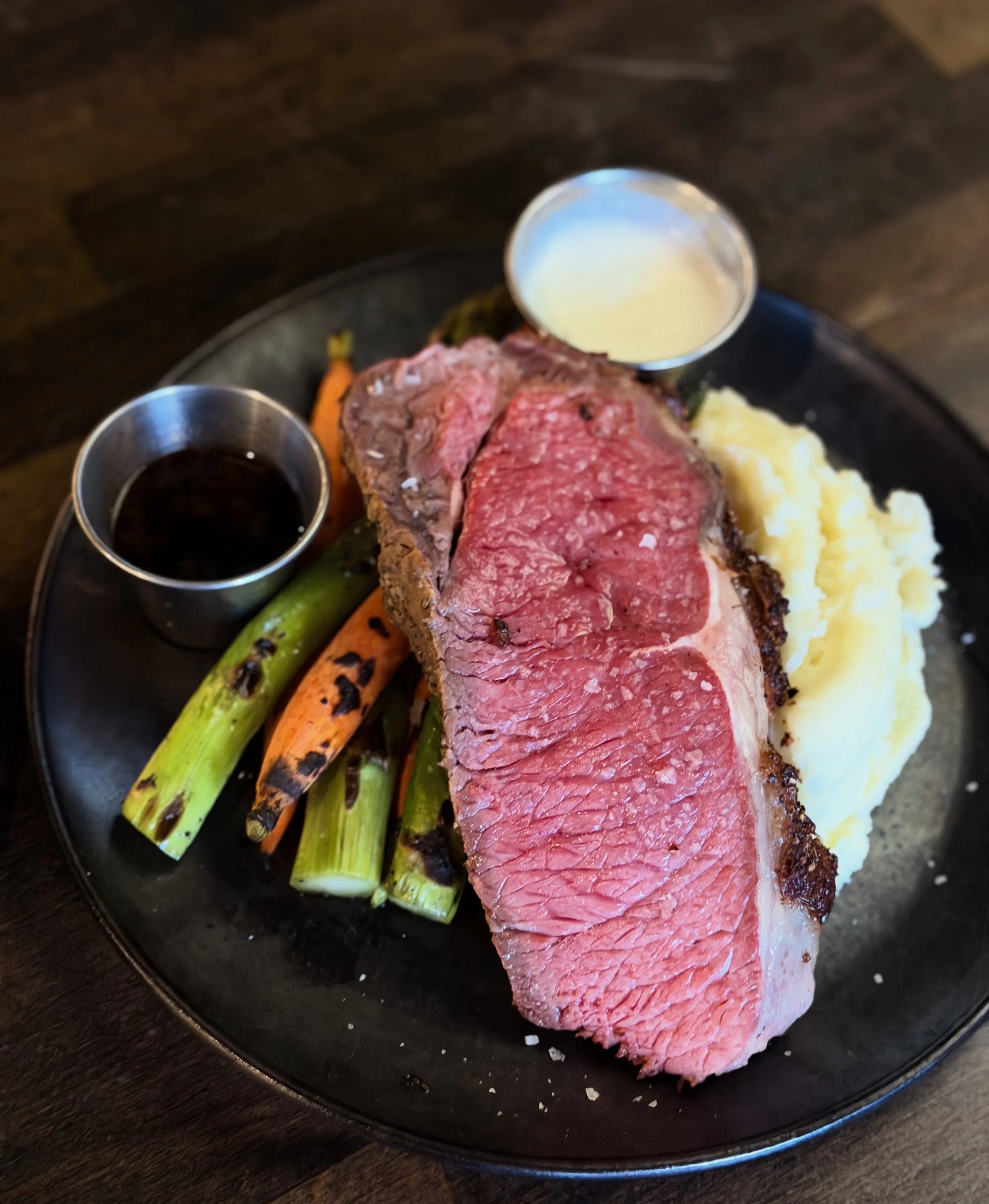 Thursday Prime Rib special; plate with a medium-rare steak, grilled carrots and green onions, mashed potatoes, and small cups of dipping sauces.