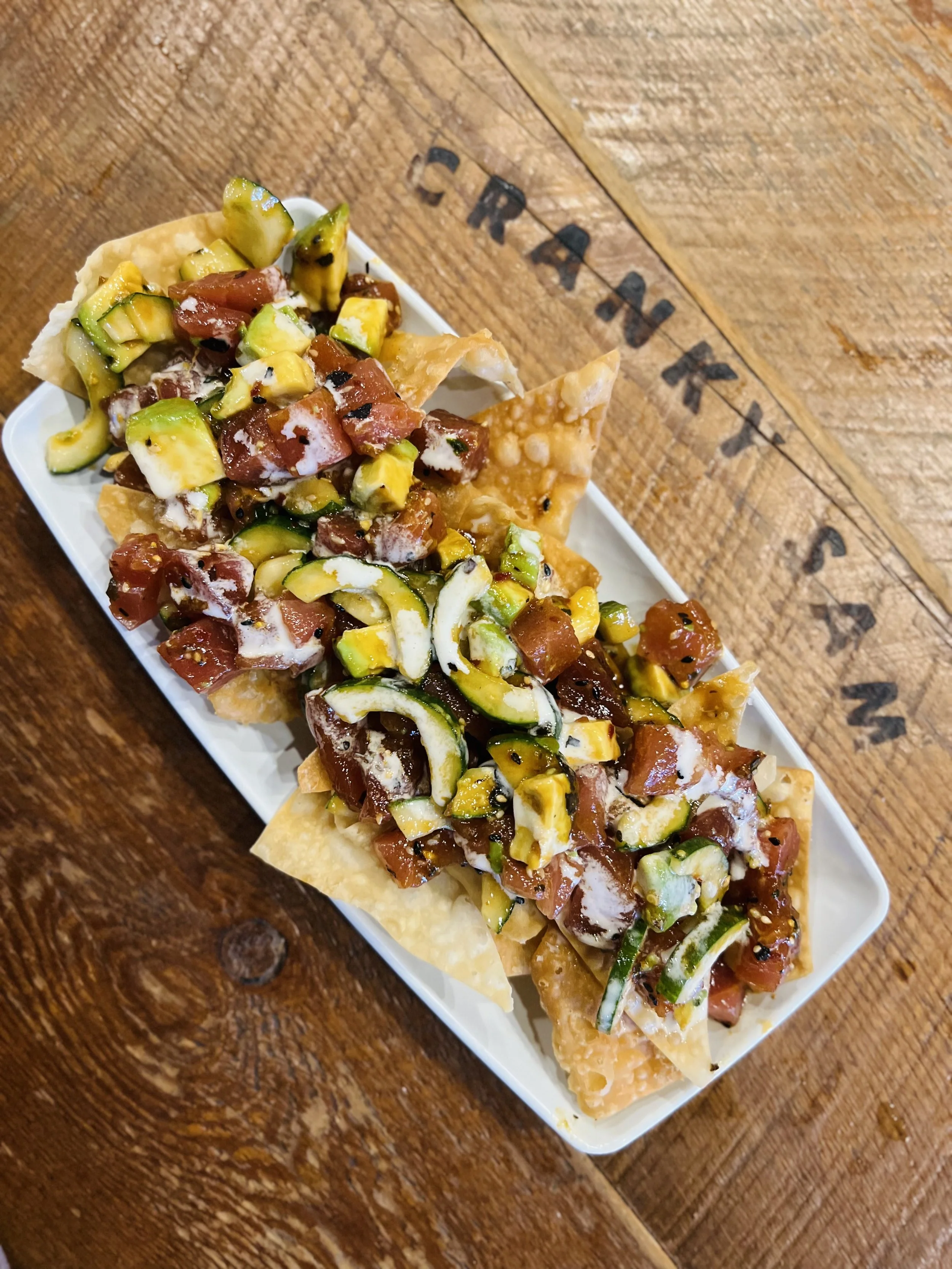 Spicy Ahi Tuna Nachos: Close-up of fried tortilla chips topped with chopped tomatoes, avocado, and herbs, served on a white rectangular plate on a wooden table.