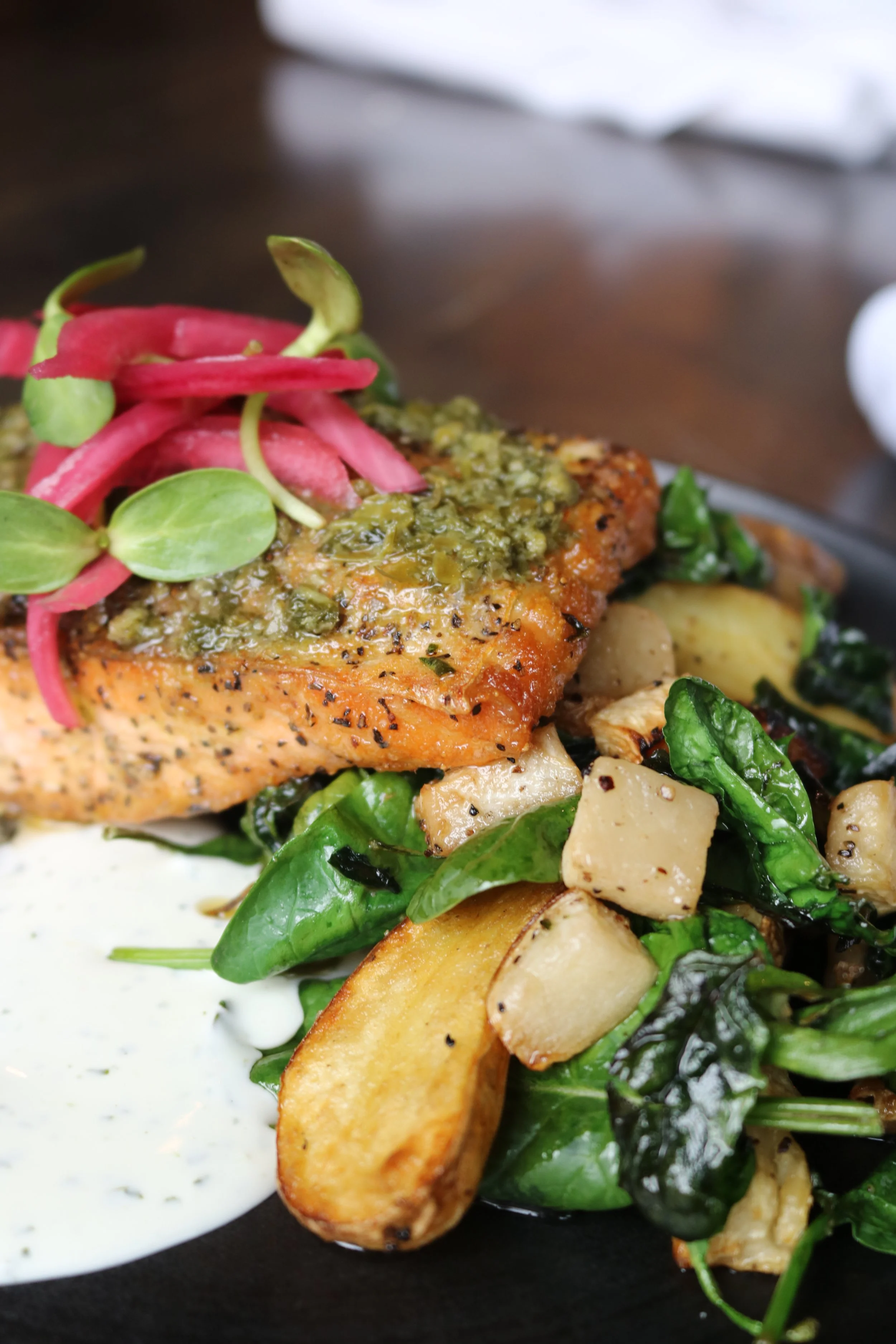 Pan Seared Salmon: A plated dish featuring a cooked salmon fillet topped with herbed sauce, served with roasted potatoes, sautéed spinach, and pickled vegetables.