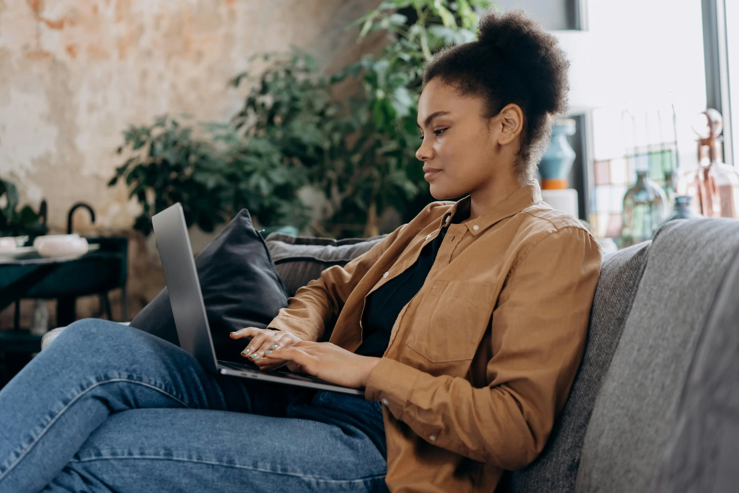Young woman sitting on a gray couch using a laptop, in a cozy living room with a stone wall, plants, and sunlight coming through a large window.