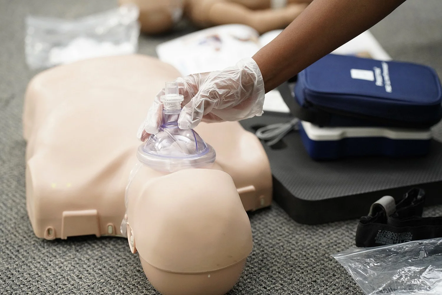 A person wearing a glove practicing CPR on a medical training mannequin, with a bag valve mask held over the mannequin's face, on a gray surface with medical supplies and equipment nearby.