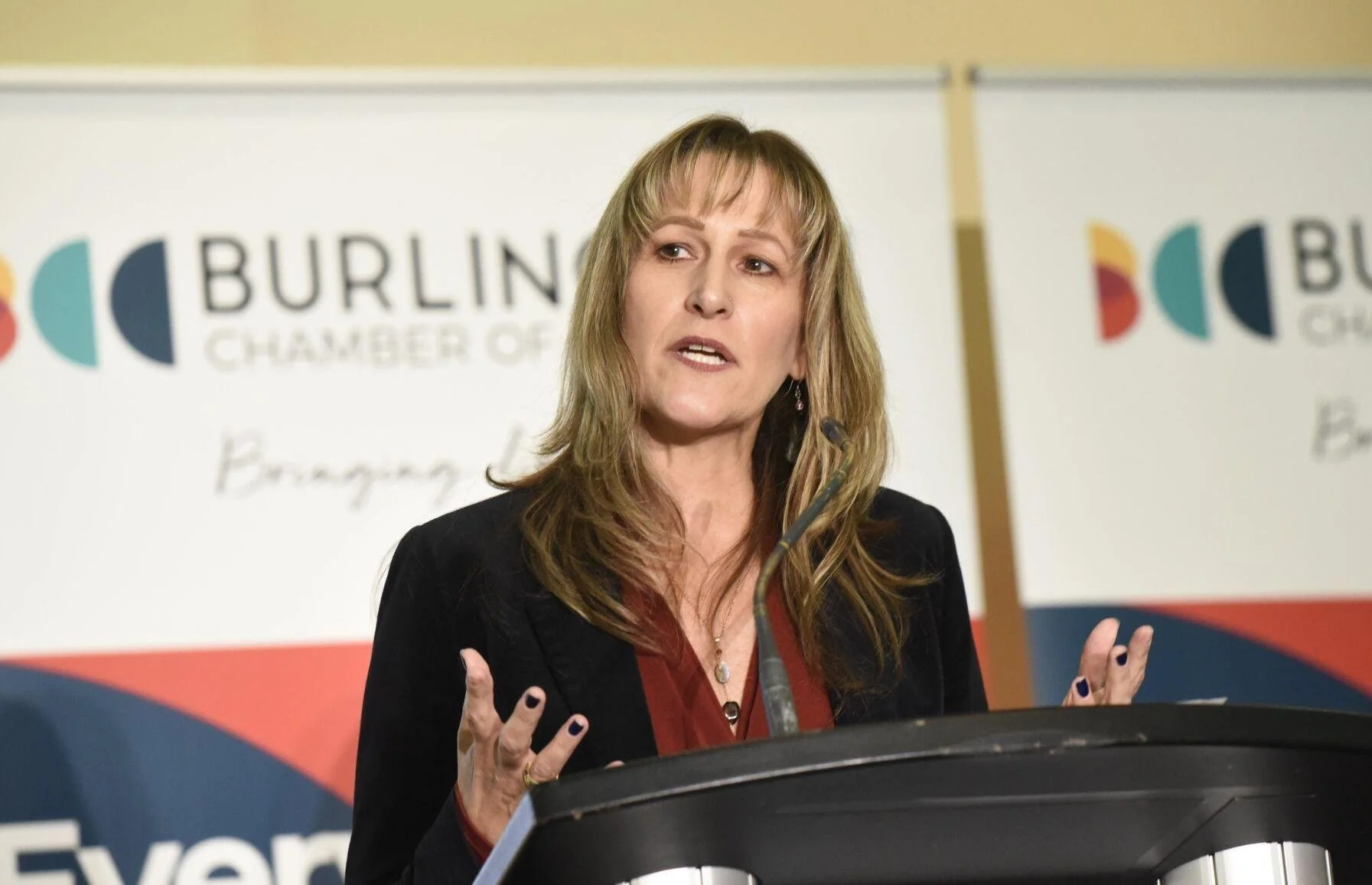 Mayor Marianne Meed Ward speaking at a podium during an event at the Burlington Chamber of Commerce, with a branded backdrop behind her.