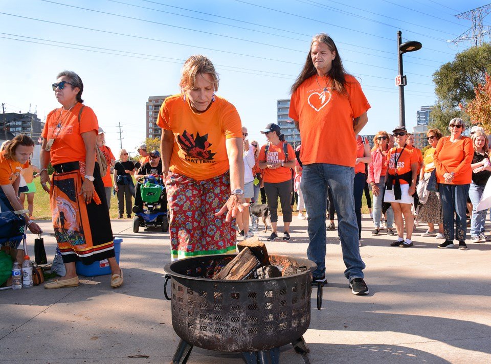 Mayor Marianne Meed Ward with a group of people gathering outdoors, wearing orange shirts, with the Mayor lighting a fire in a portable fire pit while others watch during an Indigenous-led Orange Shirt Day ceremony.