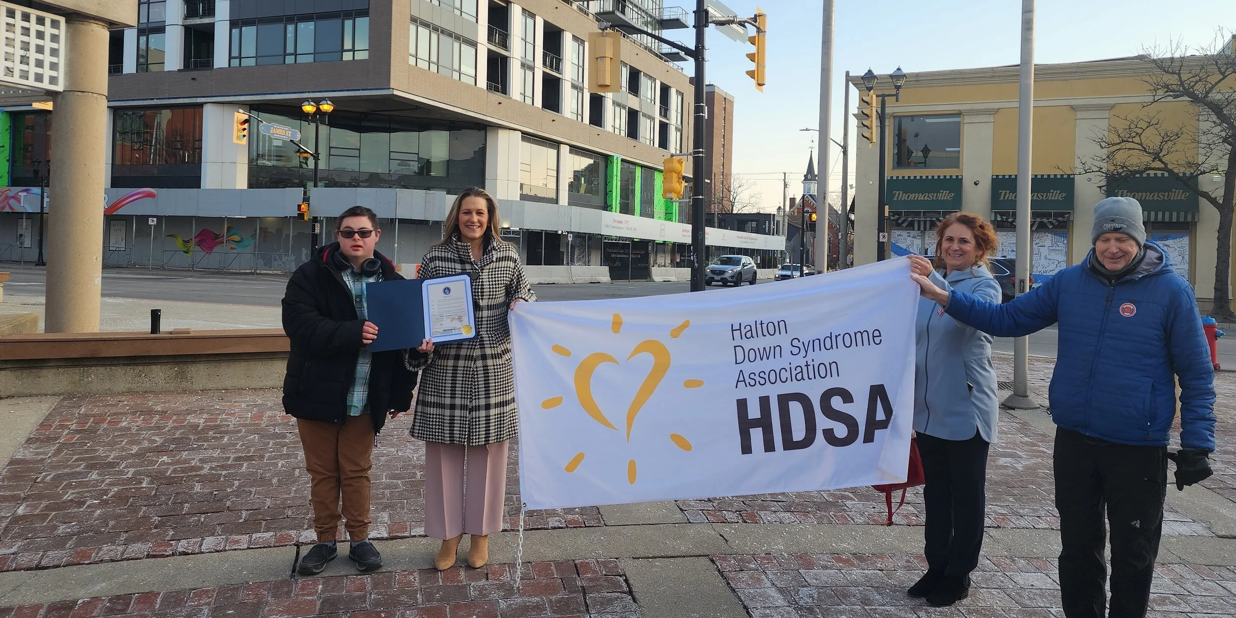 Four people, the Mayor and a women with two men, standing outdoors on a brick sidewalk, holding a banner that reads "Halton Down Syndrome Association HDSA," with a symbolic heart and sun rays design.