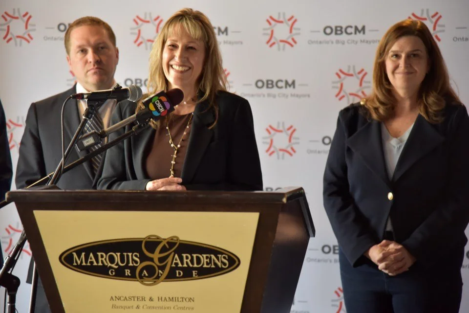 Mayor Marianne Meed Ward with one man and woman standing behind her at a podium for a press conference. The Mayor is in the centre speaking into microphones. The backdrop has the OBCM logo and the text 'Ontario's Big City Mayors.'
