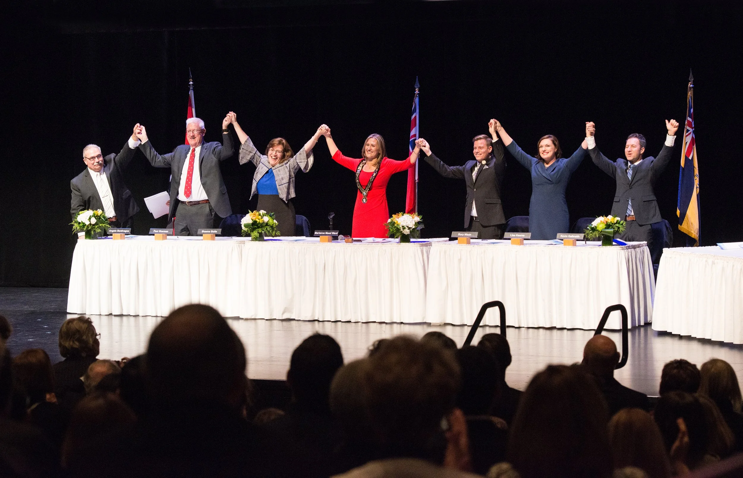 Mayor Marianne Meed Ward with newly-elected members of Council on stage holding hands in celebration at a formal event.