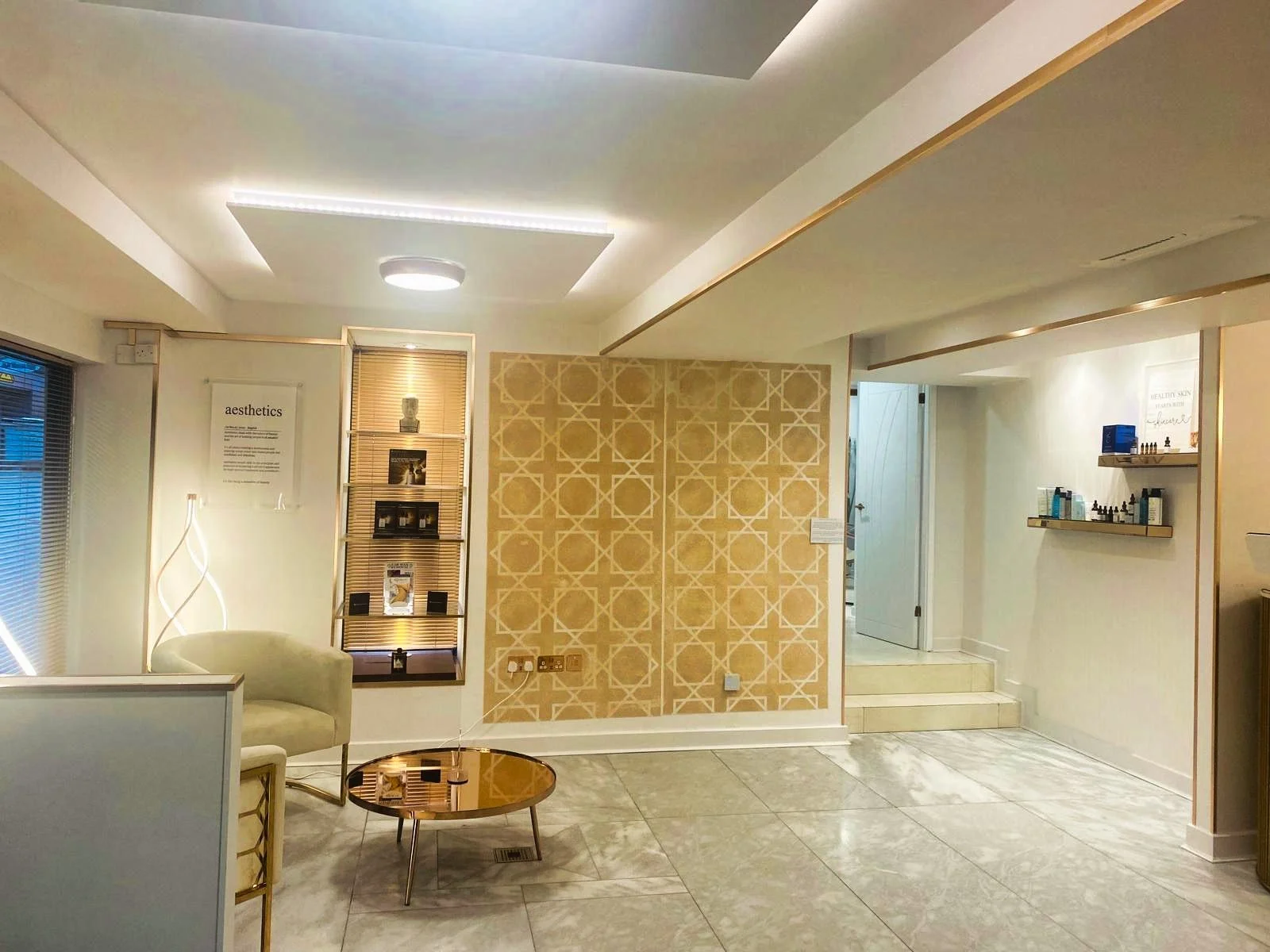 Interior of a modern spa or wellness center with decorative gold patterns on the wall, shelves with skincare products, a white armchair, a gold coffee table, and soft white lighting.