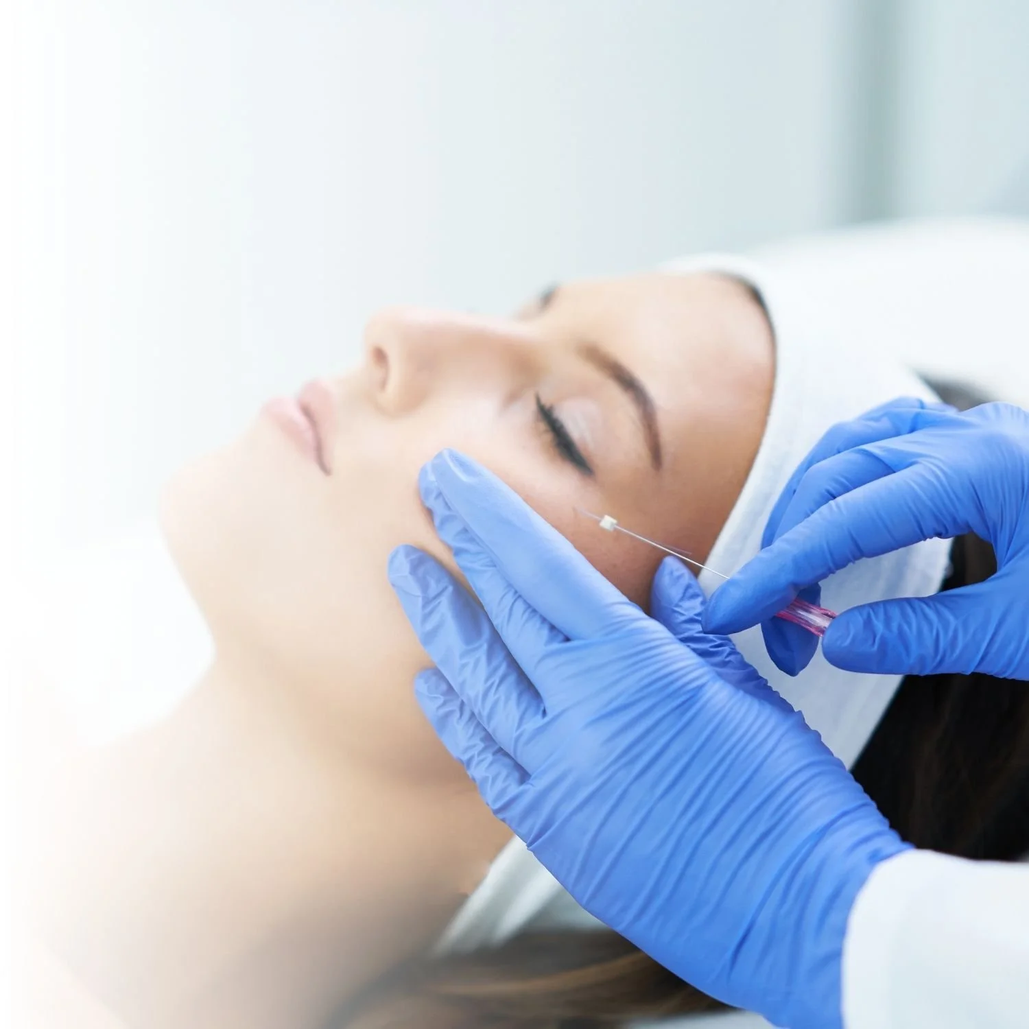 Person receiving cosmetic injection in cheek with a needle, wearing a headband, and attended by medical professional in blue gloves.