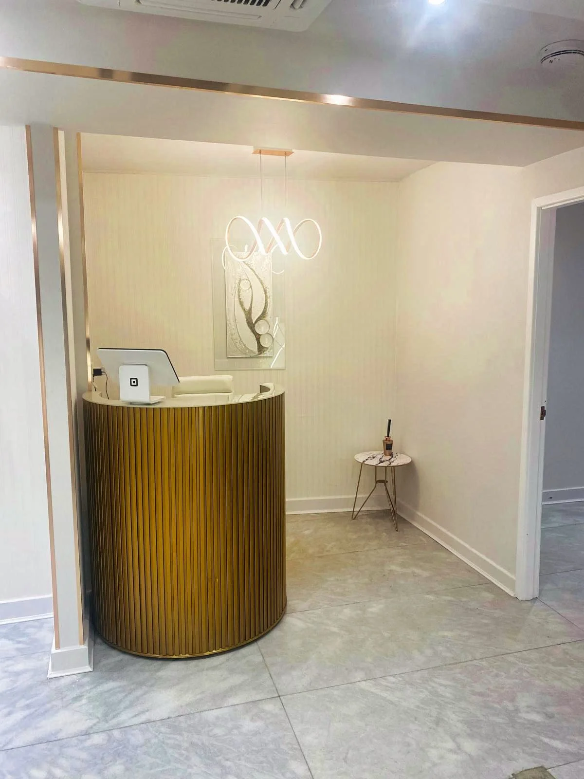 A modern reception area with a curved wooden desk, a small side table with decor, a picture on the wall, and a contemporary chandelier.