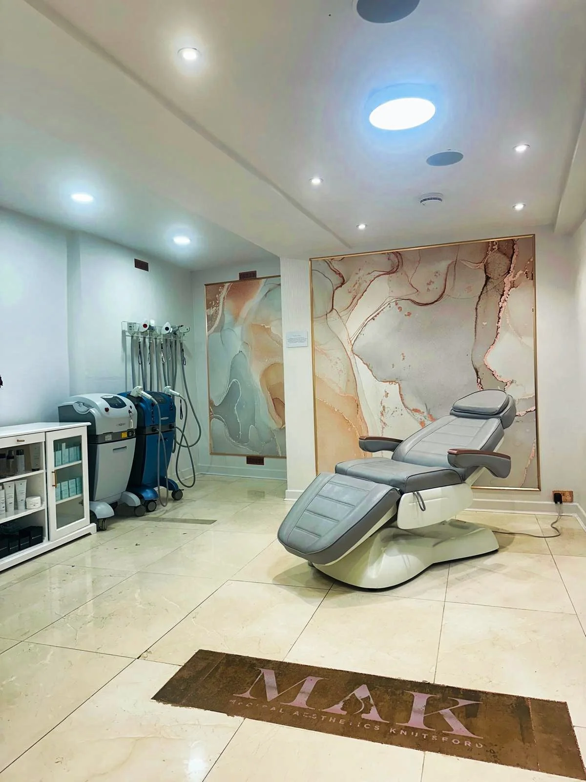 Spa treatment room with a reclining chair and medical equipment, marble wall art, and a brown mat with 'VIAIR Medical Aesthetics Knutsford' on the floor.