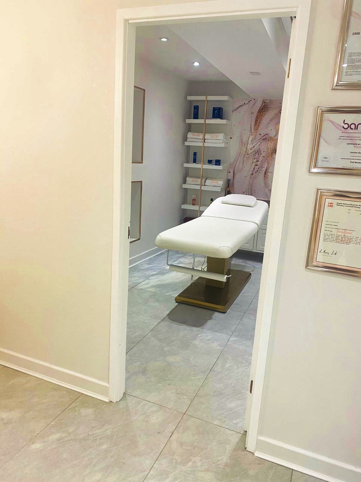 Empty medical examination room with a white adjustable treatment bed, shelves with medical supplies, and framed certificates on the wall.