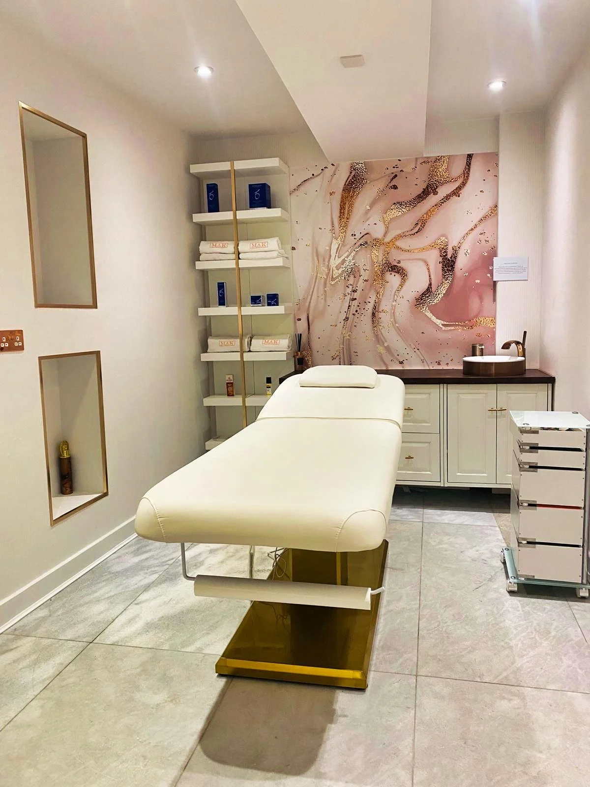 Spa treatment room with a massage bed, decorative wall art, storage shelves with supplies, and a small sink area.