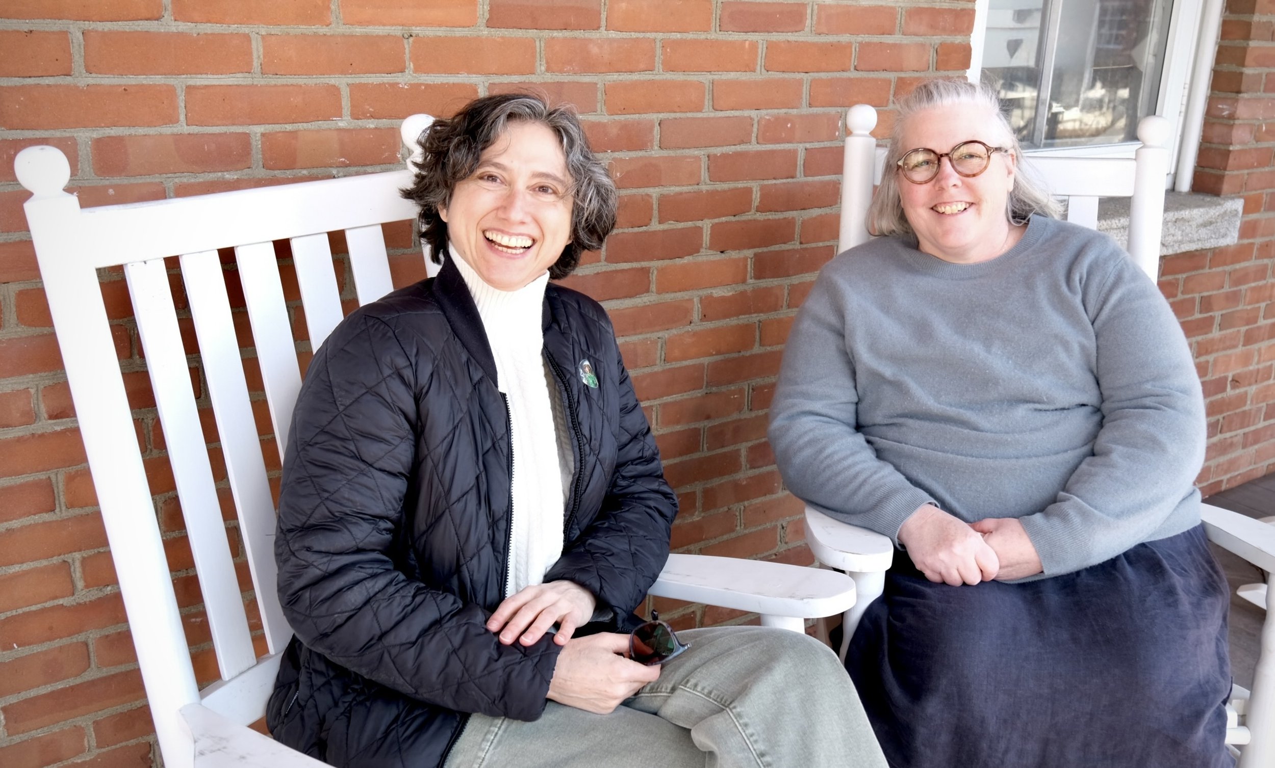 Miranda Jane and Amy Noyes sitting on white rocking chairs outside, smiling, against a brick wall in Montpelier, VT. One woman has gray hair and glasses, wearing a gray sweater and dark skirt. The other woman has curly dark hair..