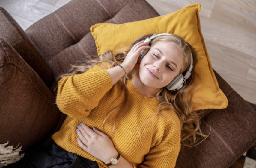 A white, autistic teenage girl with blonde hair lies on a brown couch. Her head is on a yellow pillow. She is wearing headphones. Her eyes are closed and she has a soft smile on her face. Her hand is resting on her stomach.