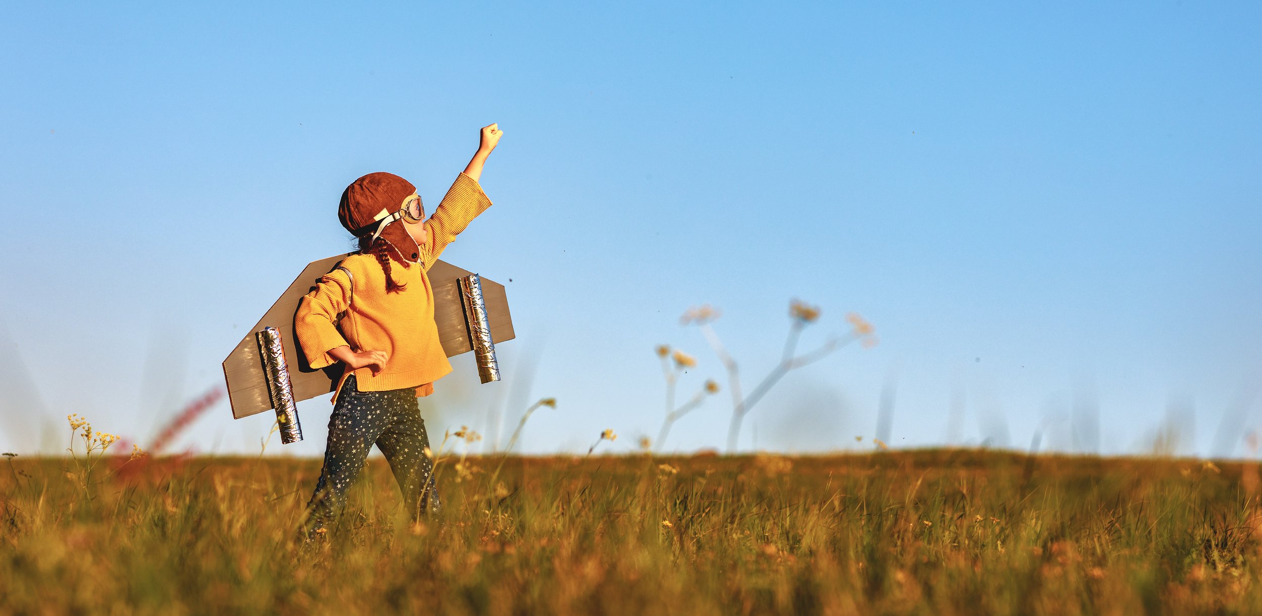 An autistic child dressed as a rocket with a cardboard body and aluminum foil engines, standing in a grassy field with wildflowers, raising one arm in the air against a clear blue sky.. Neurodivergent child, ADHD, autism, autistic. Support.