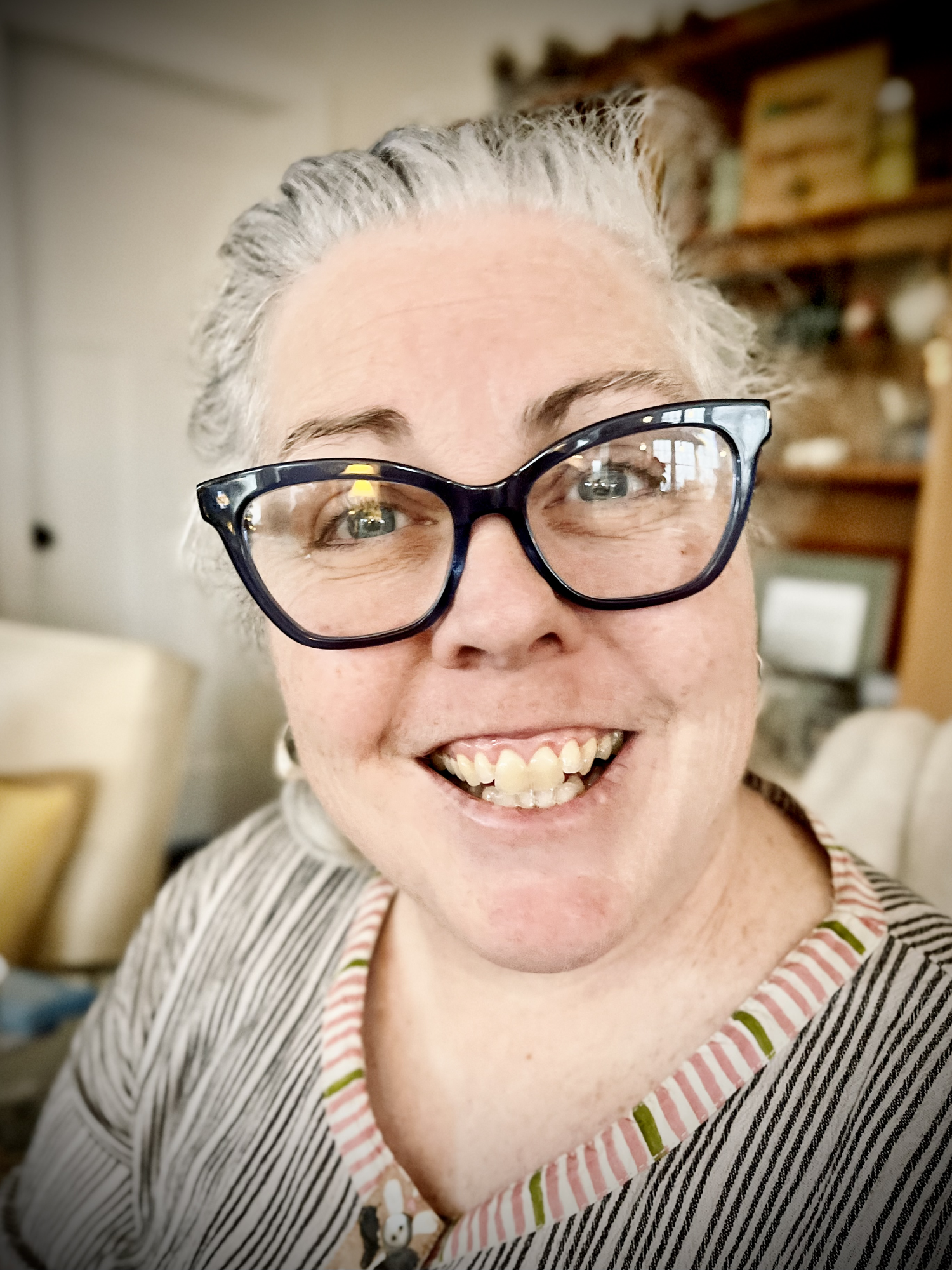 Close-up of Amy Noyes with hair pulled back in a ponytail, wearing large black glasses and a striped shirt, in a cozy home setting with wooden shelves in the background.