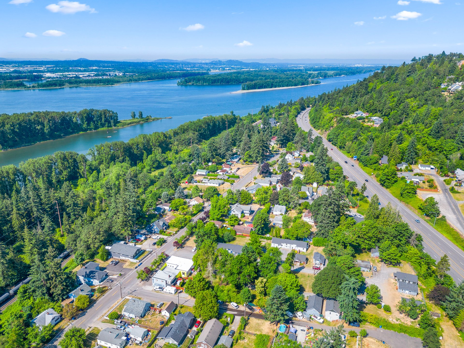 Aerial view of a small residential neighborhood by a river with lush green trees and hills in the background under a blue sky.