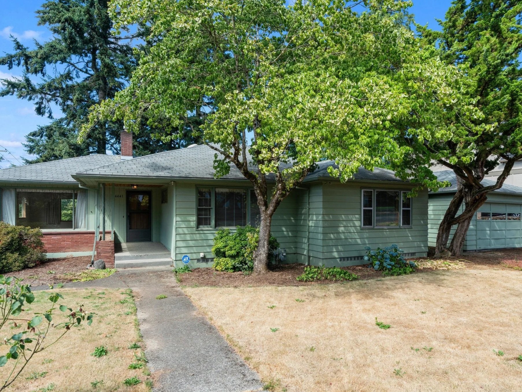 A mid-sized, single-story house with light green exterior and white trim, surrounded by trees and a front yard with a concrete walkway, with a large leafy tree in the front yard. The porch has two steps and a front door with a glass window.