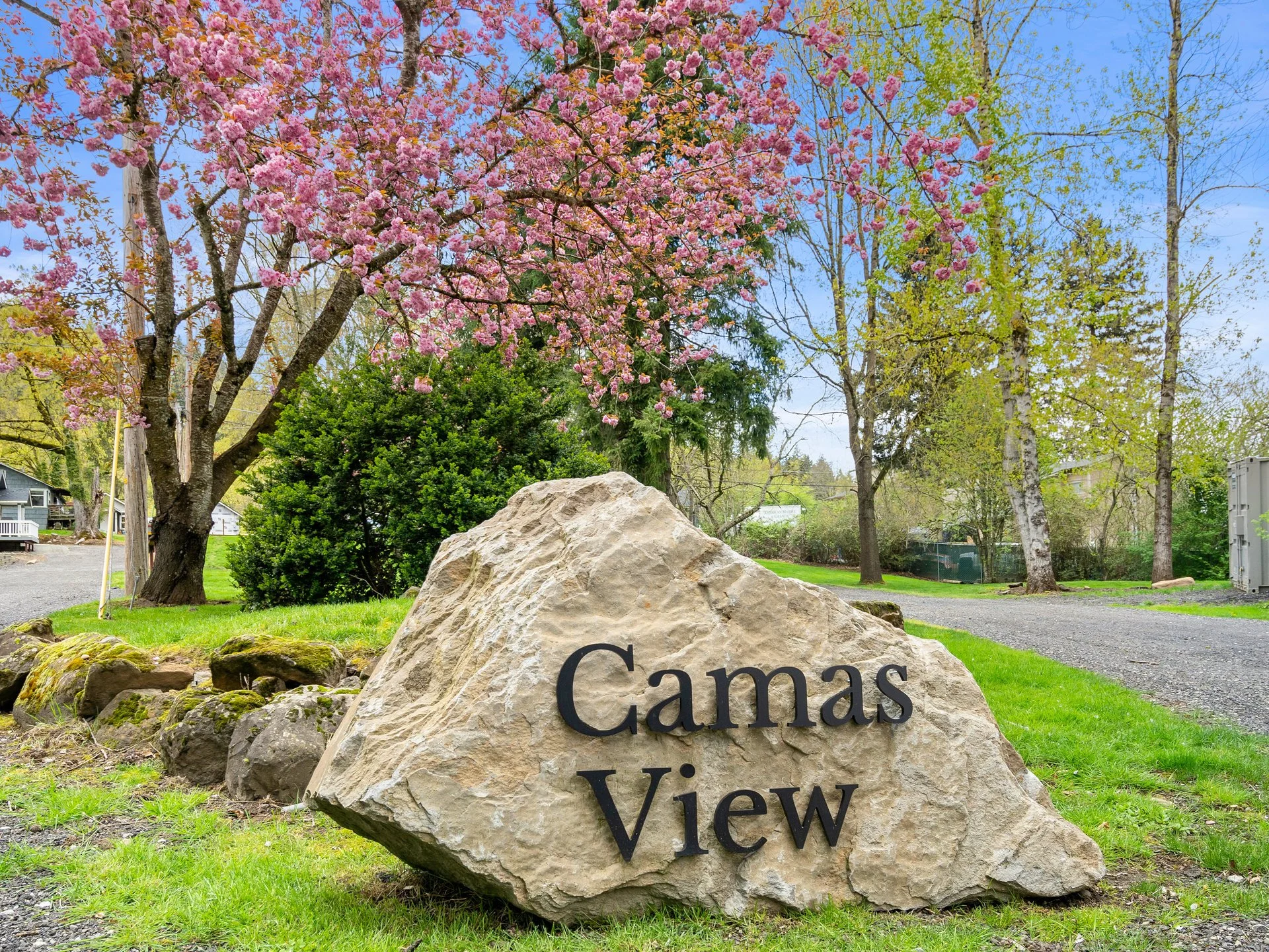 A large rock with the words "Camas View" engraved on it in front of a landscaped yard with trees, including a pink flowering cherry blossom tree, and a gravel pathway.