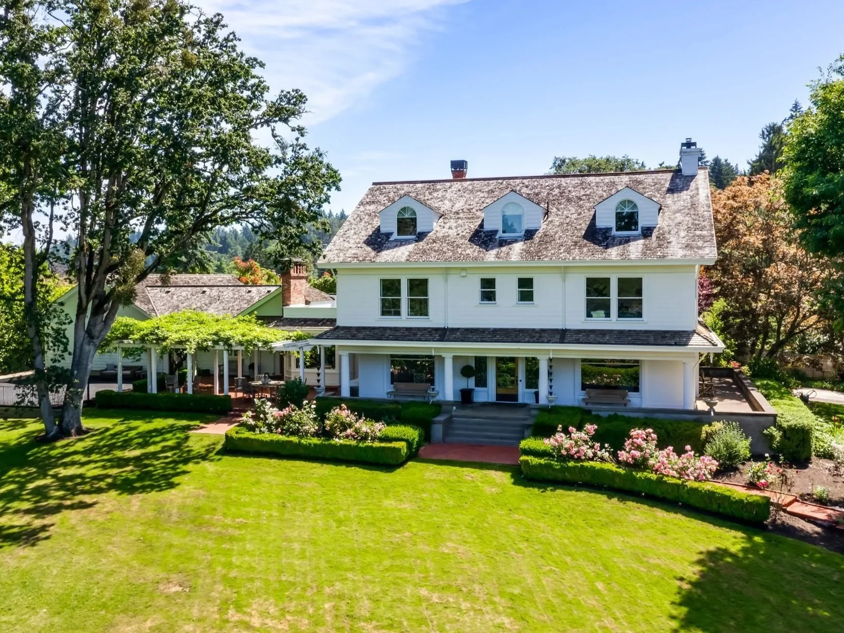 A large white house with multiple dormer windows on the roof, surrounded by a well-maintained lawn and garden, with mature trees and a patio area on the side.