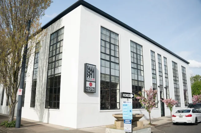 Sportslab located in the Oregon Square Office Park near Lloyd Center. White multi-story building with large black-framed windows, a sign reading 'Athletic Republic,' and pink flowering trees outside, parking lot with cars in front.