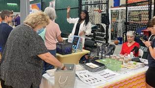 A group of people gathered around a booth at an event, with a woman explaining something and others looking at items on display, including jewelry and crafts.