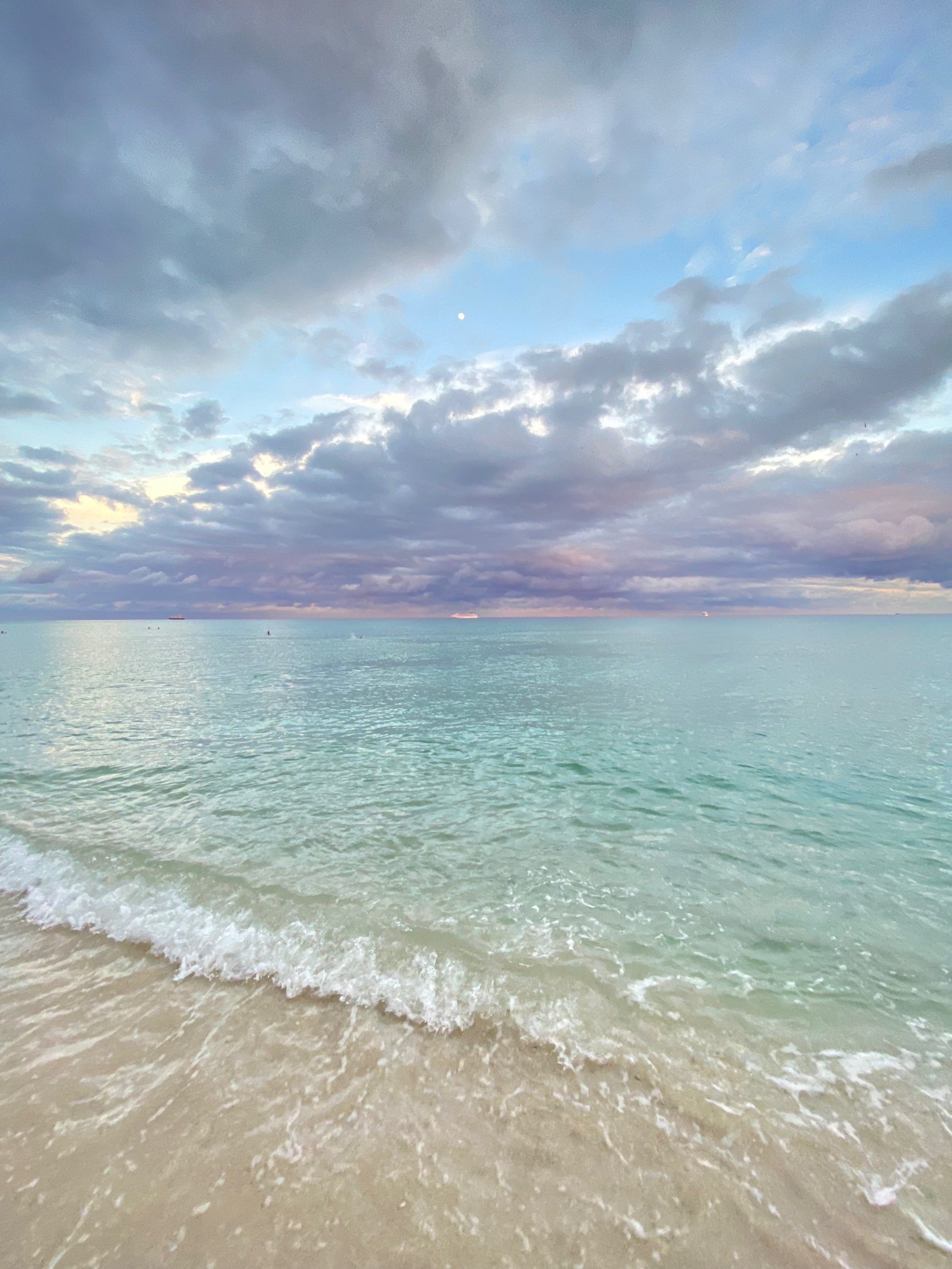 A beach scene with clear turquoise water, gentle waves on the sandy shore, partly cloudy sky, and small ships in the distance.