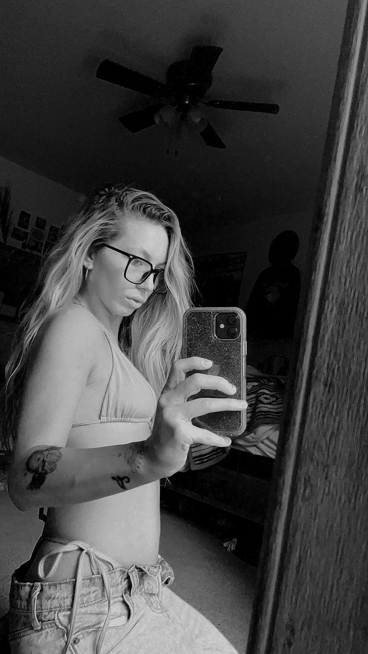 A black and white selfie of a young woman with glasses taking a mirror photo. She has long, wavy hair, tattoos on her arm, and is wearing a crop top and shorts. She is in a room with a ceiling fan and various items on the walls and in the background.