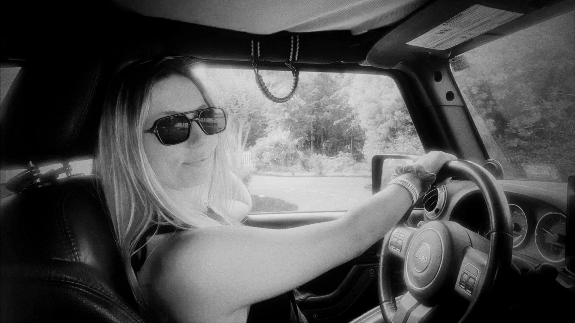 A woman sitting in the driver's seat of a Jeep, wearing sunglasses and a white t-shirt, while driving down a tree-lined road on a sunny day.
