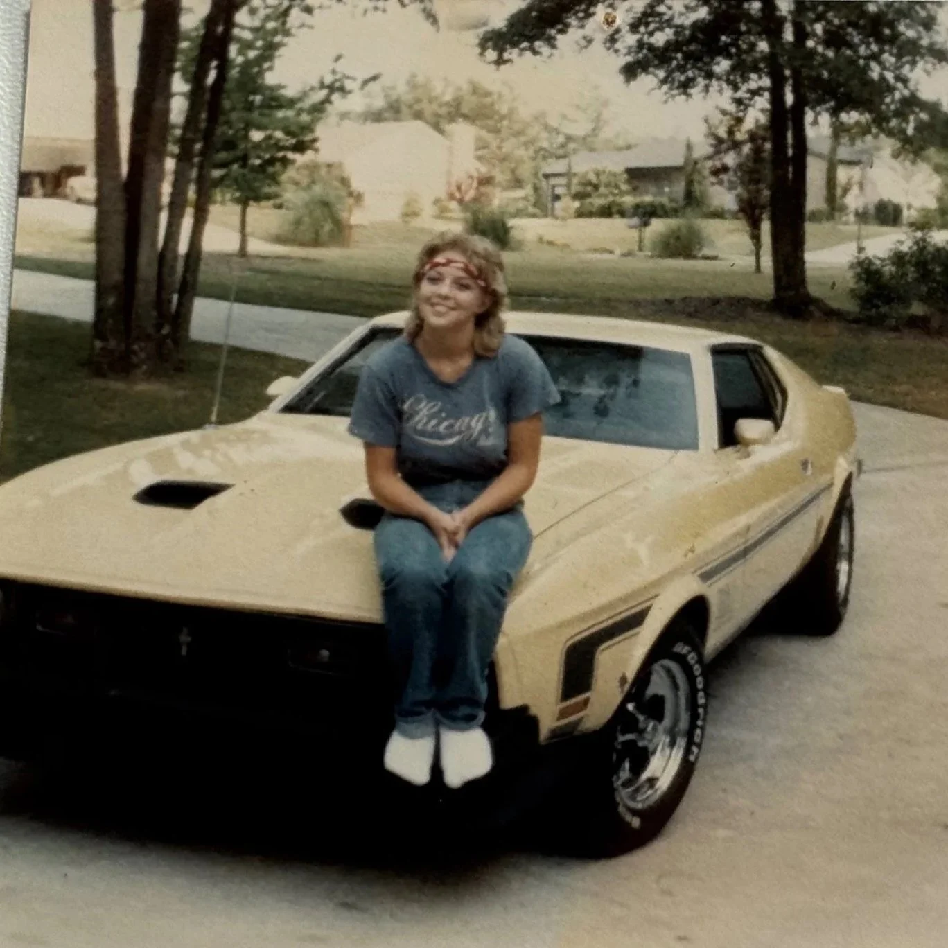 A woman sitting on the hood of a yellow vintage sports car parked on a suburban street with trees and houses in the background.