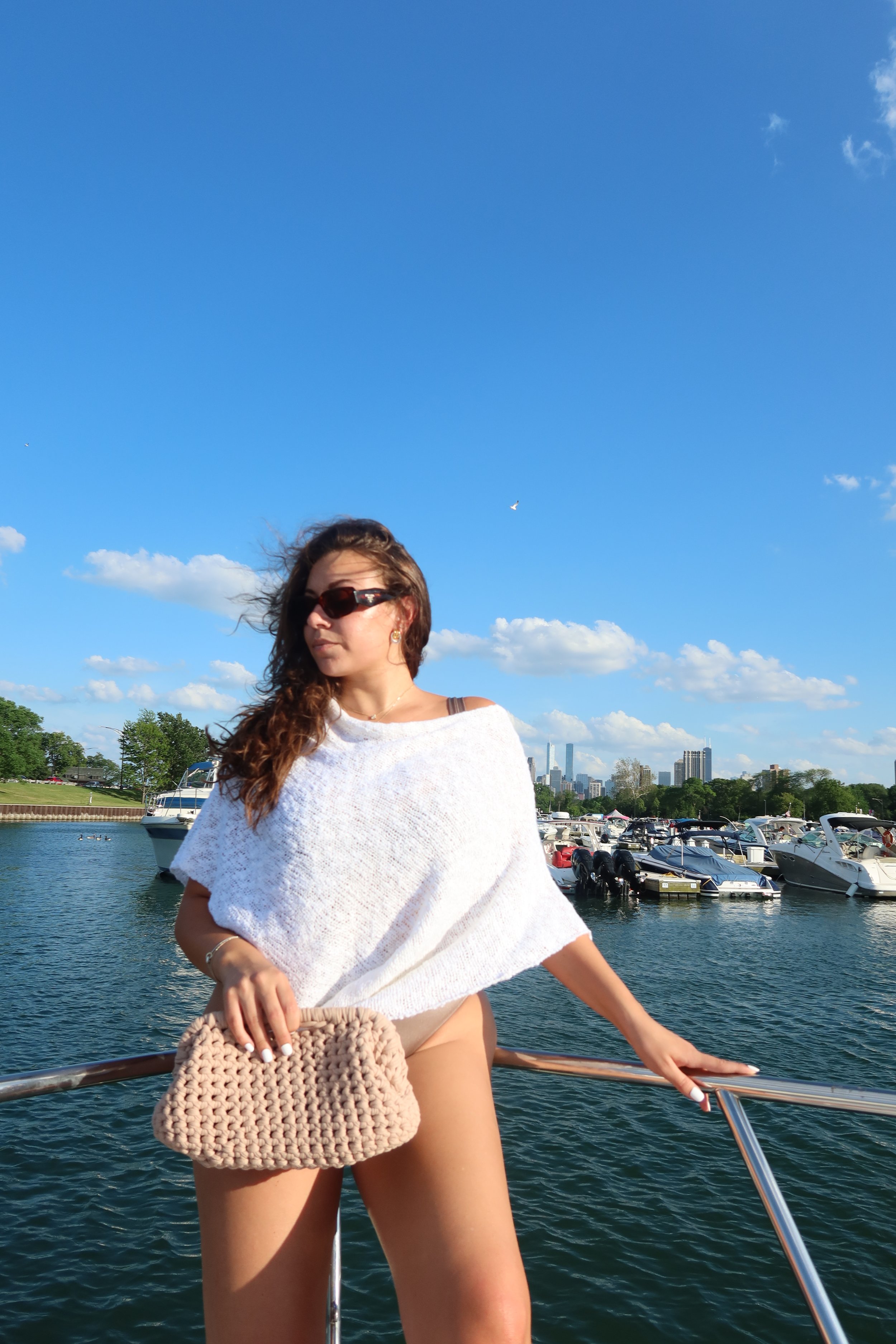 A woman with long brown hair, wearing sunglasses and a white shawl, standing on a boat with a marina and city skyline with tall buildings in the background under a blue sky with some clouds.