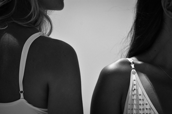 Close-up of two women standing side by side, wearing tank tops, with one woman’s shoulder and part of her hair visible on the left, and the other's shoulder and hair on the right.