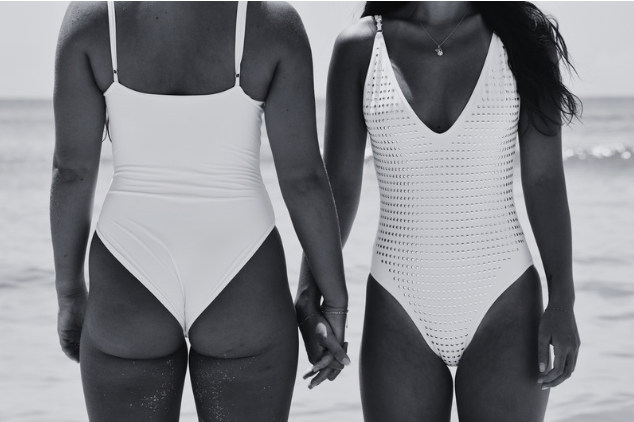 Two women in swimsuits holding hands on the beach, seen from behind.