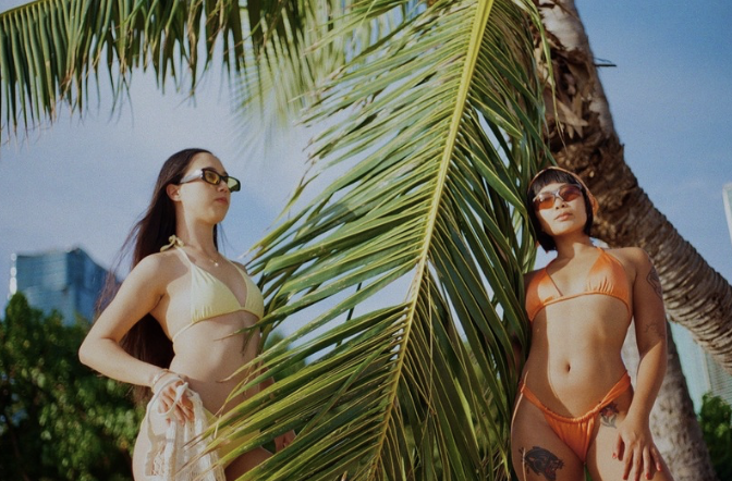 Two women in swimsuits standing among palm trees outdoors on a sunny day.