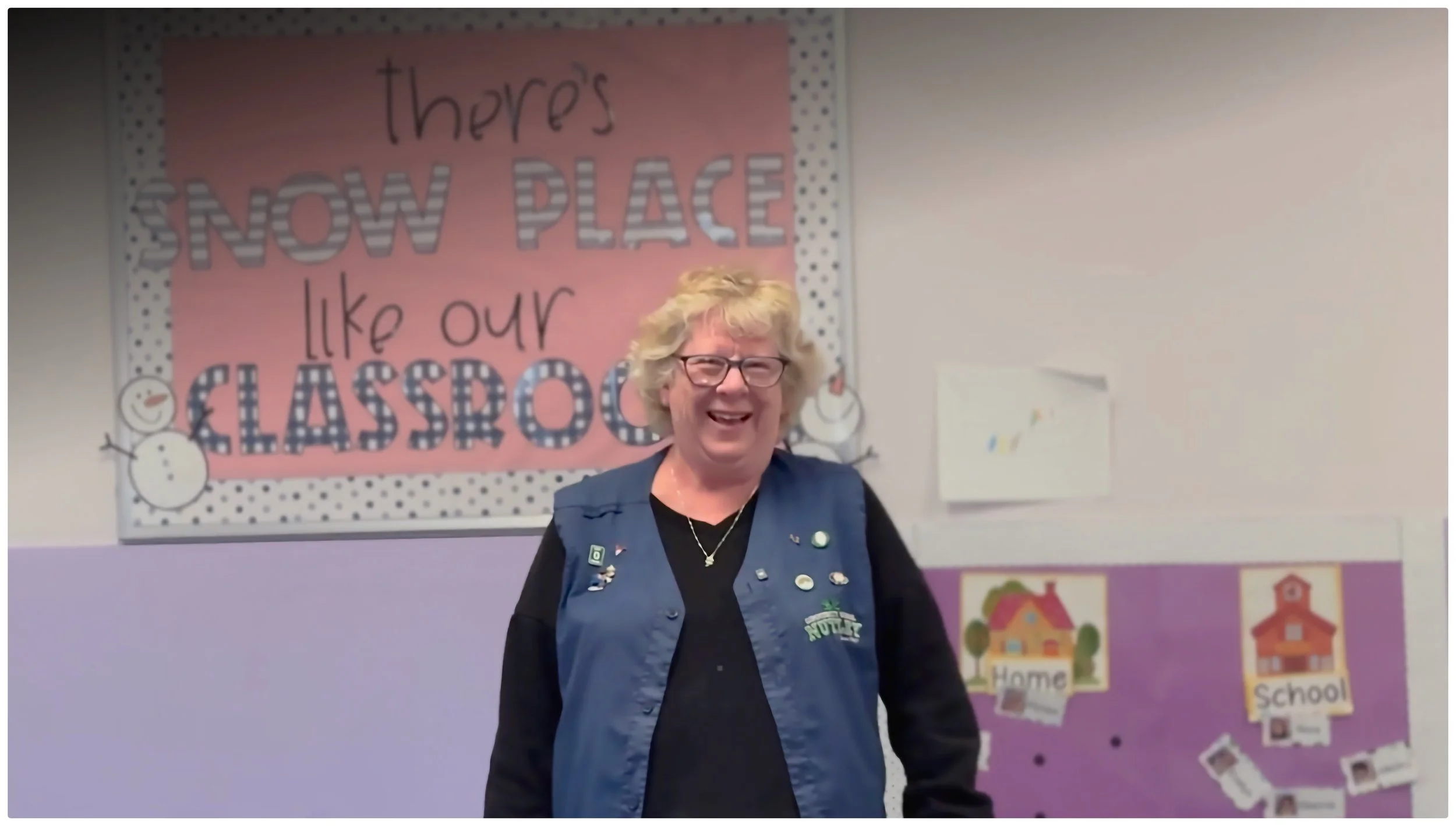 A smiling woman with glasses and blonde curly hair standing in front of a classroom bulletin board that reads 'there's no place like our classroom' with a snowman drawing. The bulletin board has pink and purple sections with school-related decorations.