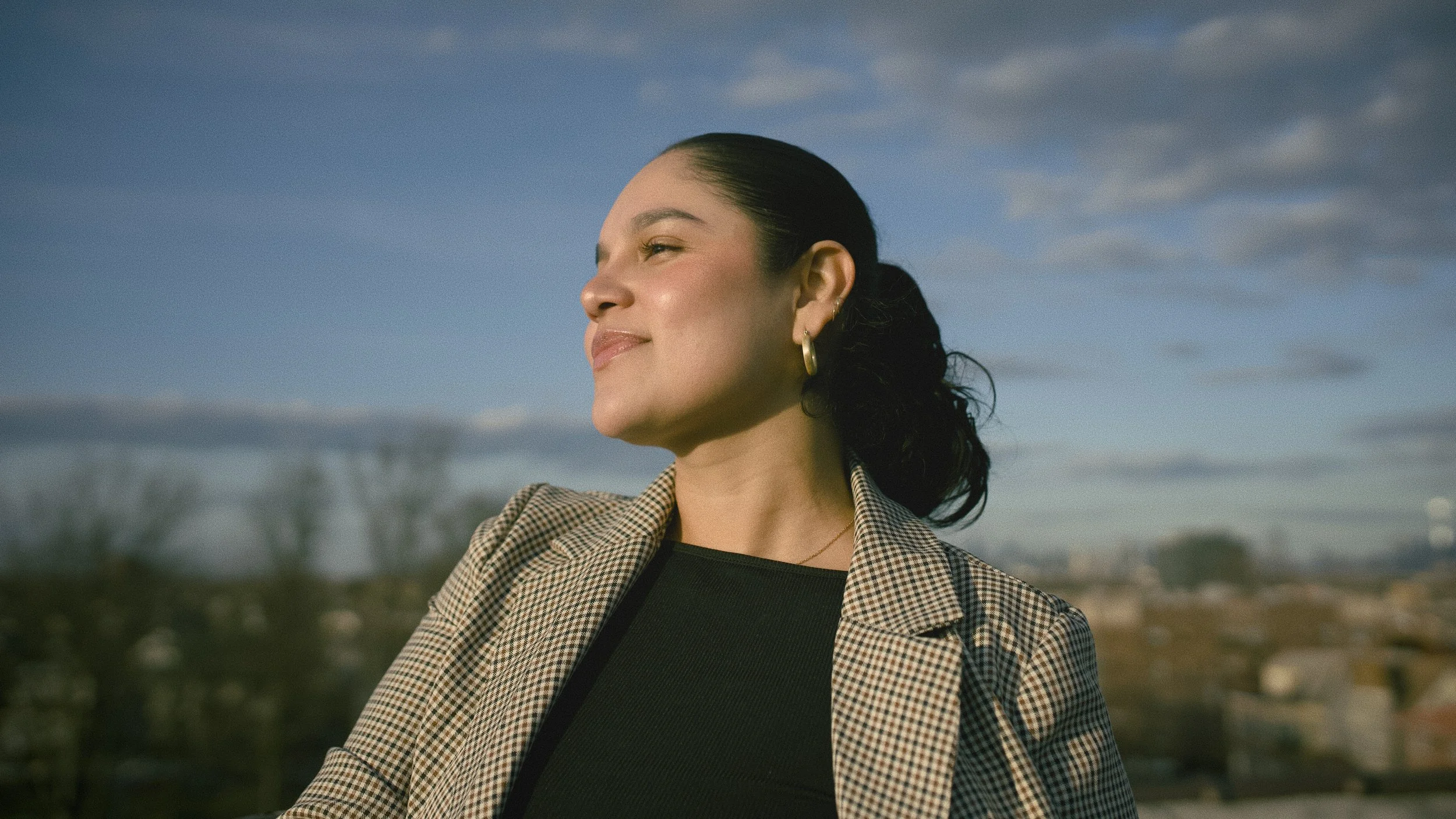Side profile of a woman with dark hair pulled back, smiling with her eyes closed, standing outdoors during daytime with a cloudy sky and cityscape in the background in Bayonne New Jersey.