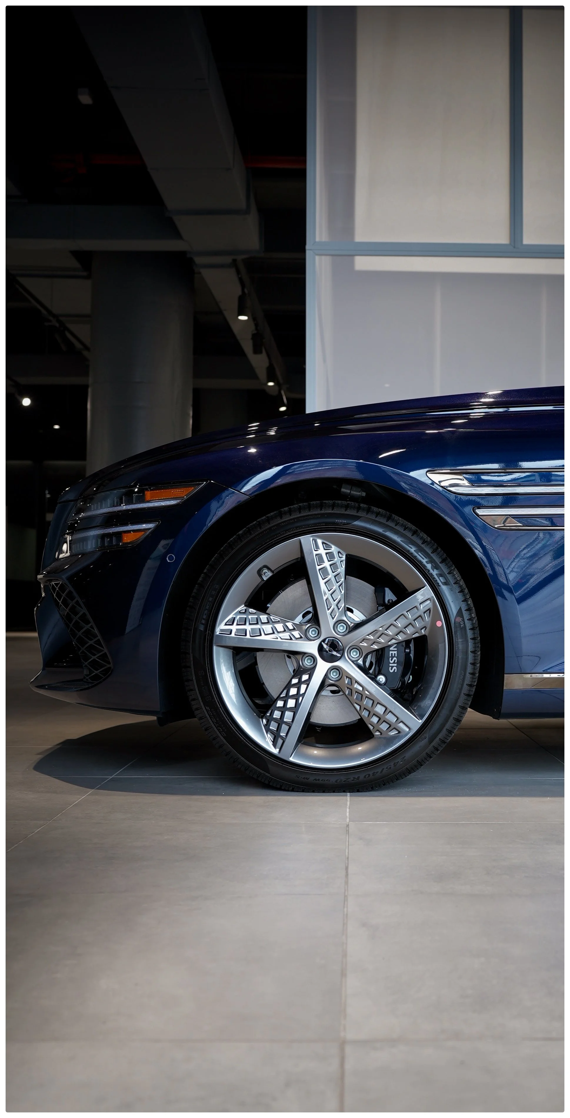 Close-up of the front left side of a dark blue Genesis g80 model car in a Brooklyn showroom located in New York, showing the wheel and part of the car's body.