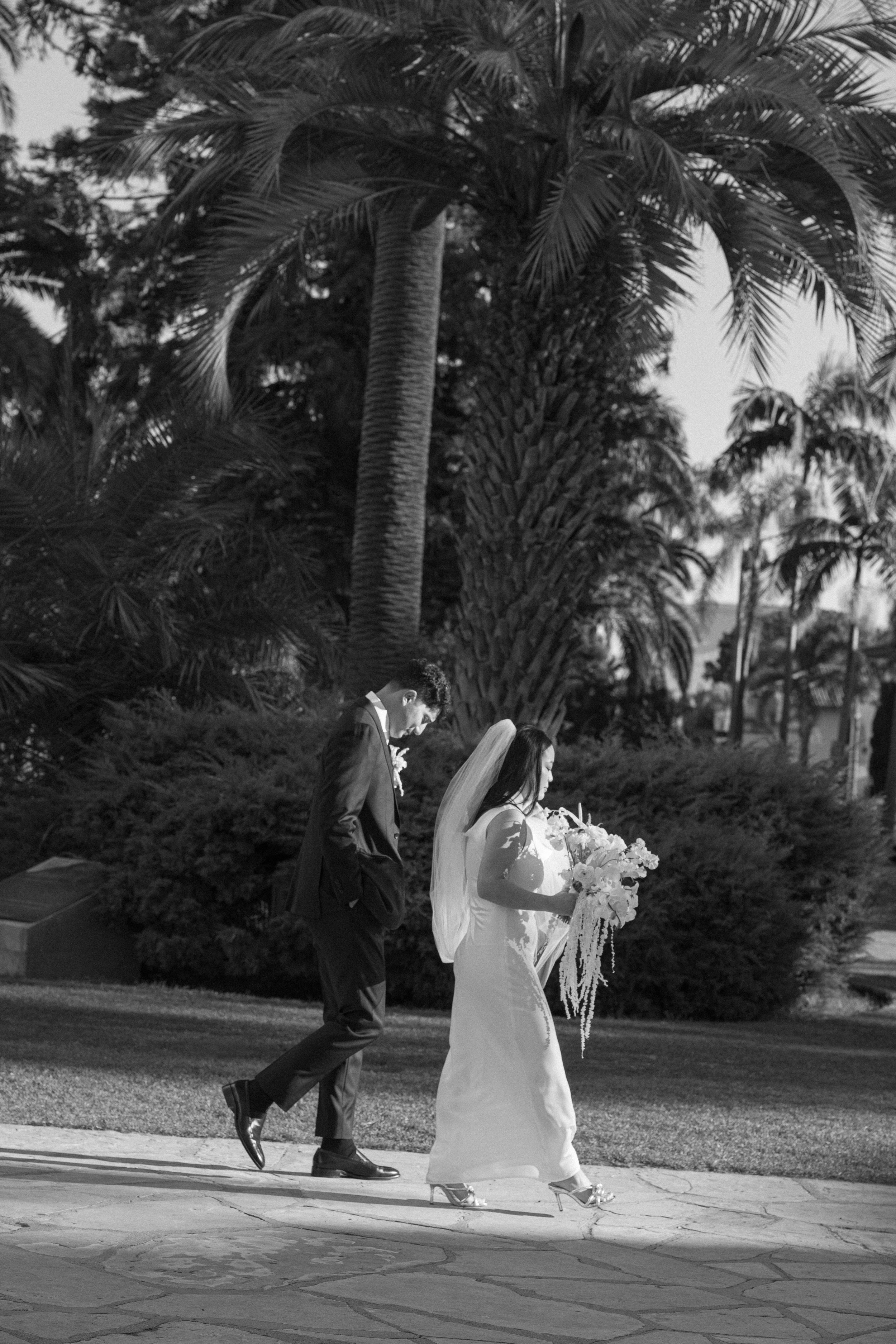 Black and white photo of a bride and groom walking side by side with palm trees in the background.