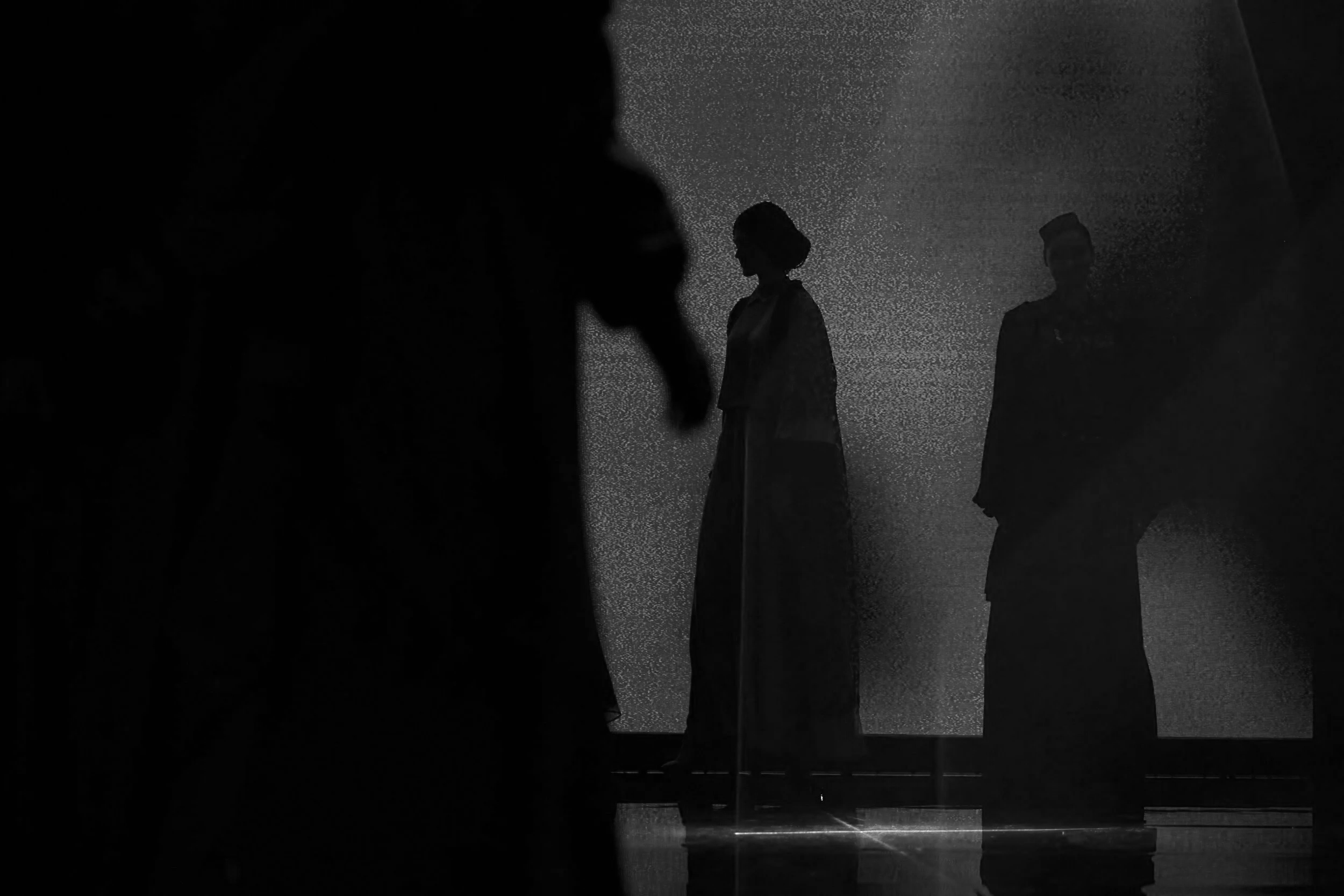Silhouettes of people standing and walking indoors, with a textured background and a vertical support beam.